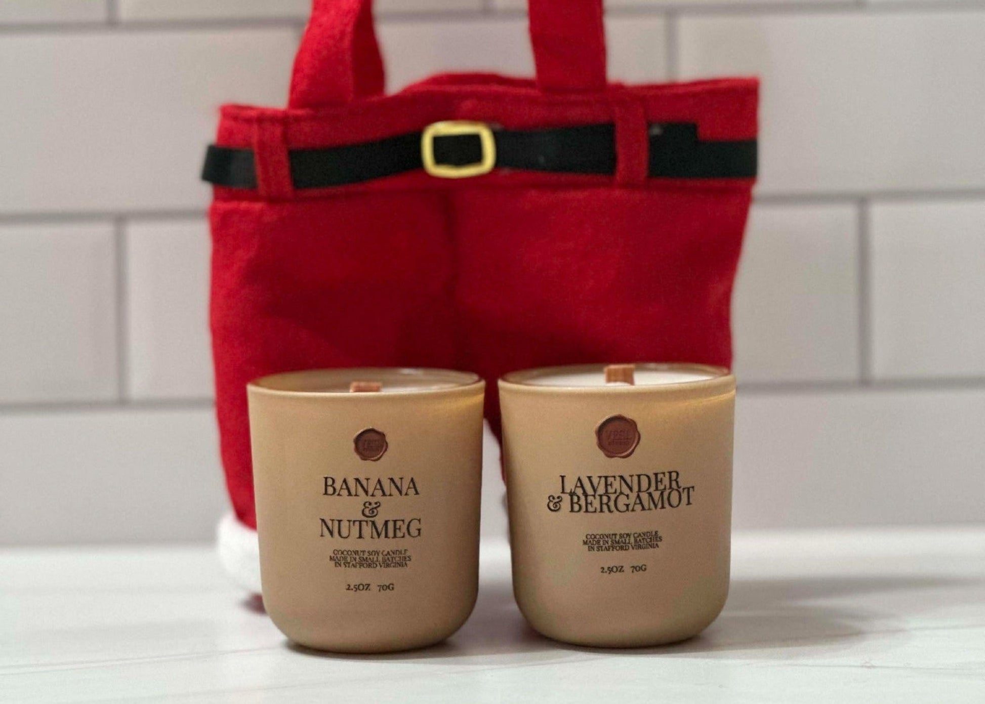 Two mini candles labeled 'Banana & Nutmeg' and 'Lavender & Bergamot' in front of a red gift santa pants treat bag with a belt buckle.