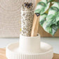 White ceramic holder with smoldering sage sticks on a wooden surface with a plant in the background