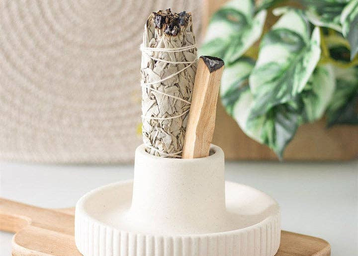 White ceramic holder with smoldering sage sticks on a wooden surface with a plant in the background